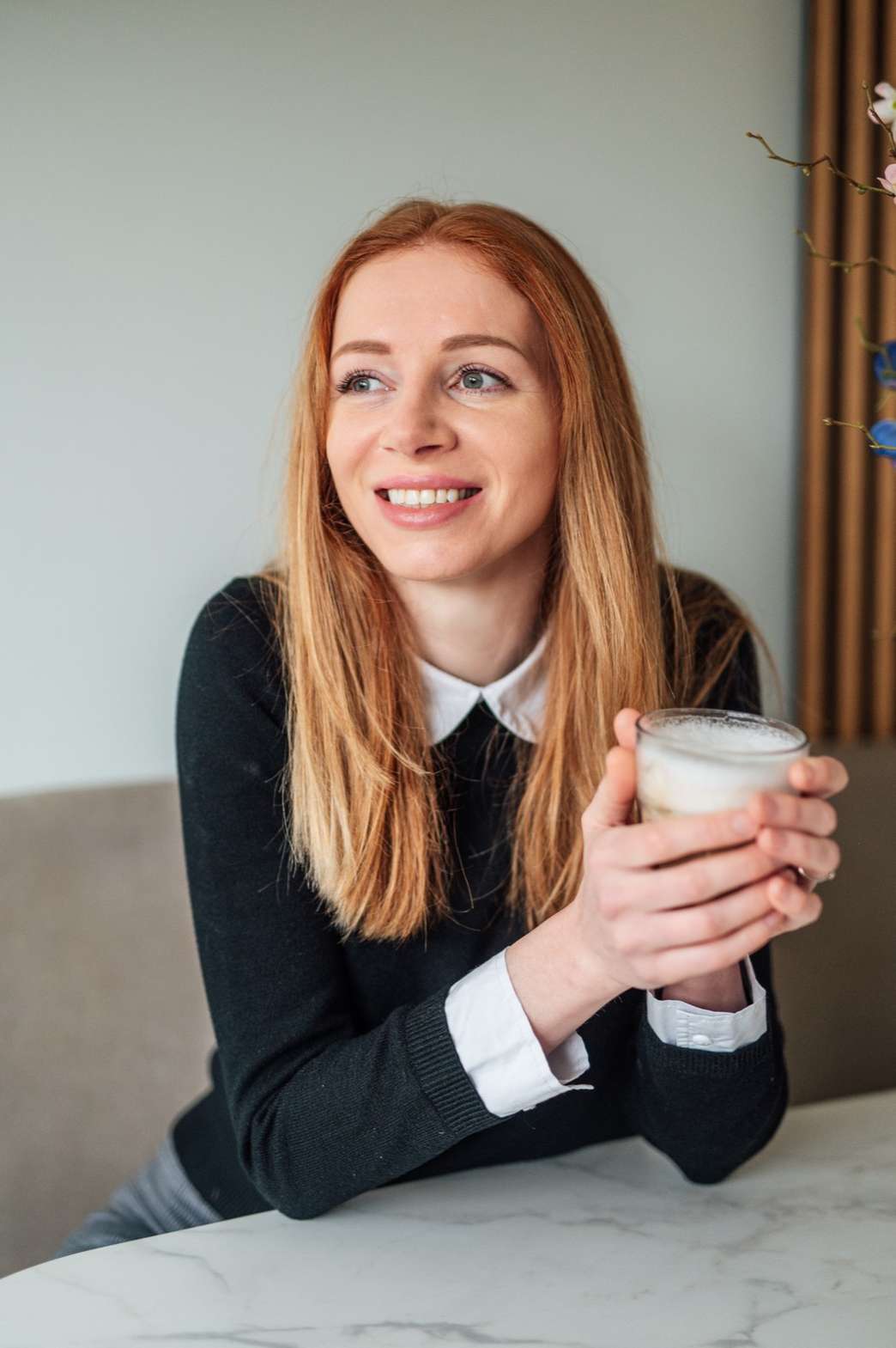 Marjolein Ligtvoet aan tafel met een glas koffie in haar handen, glimlachend en opzij kijkend.