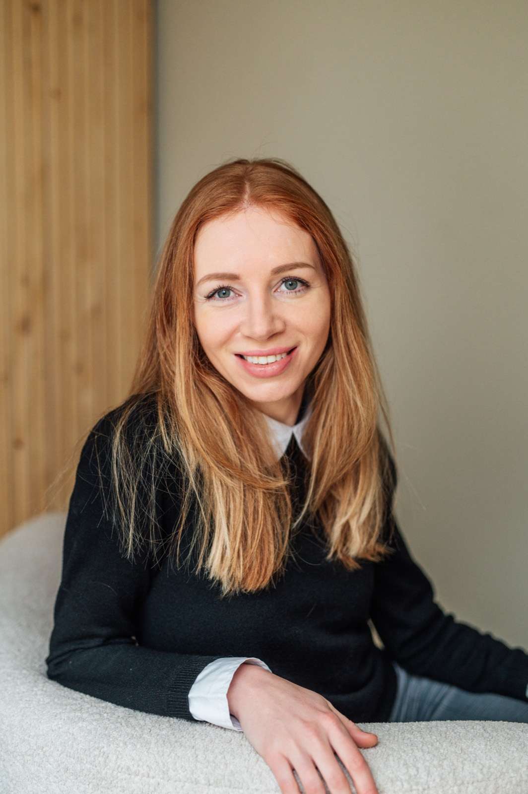 Marjolein Ligtvoet, zittend in een zwarte trui met witte kraag, lacht in de camera. Achter haar een wand met eikenhouten lamellen.