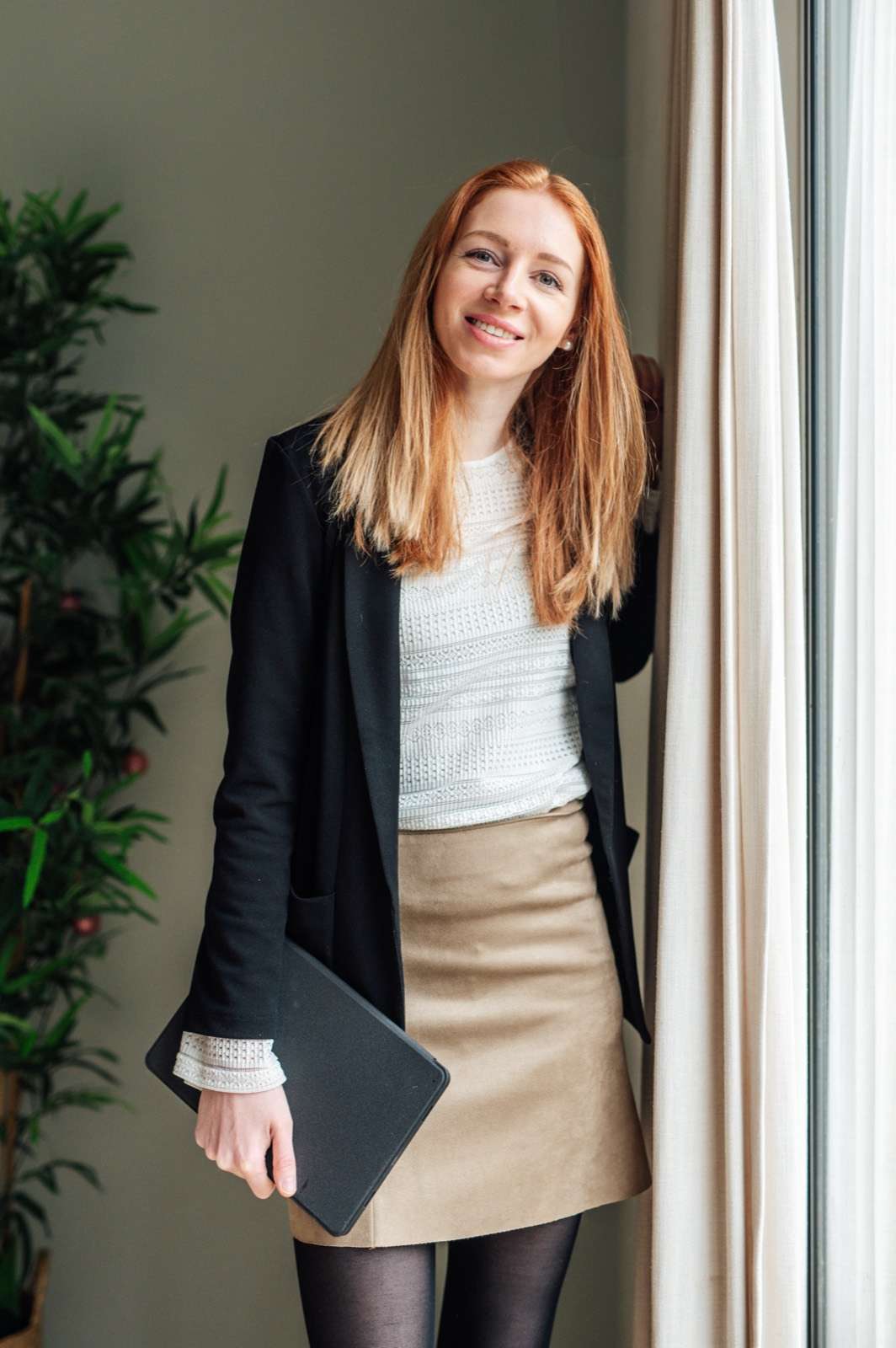 Marjolein Ligtvoet staat bij een raam met lichte gordijnen, in een zwart colbert en een beige rok, met een map onder haar arm. Ze lacht in de camera.